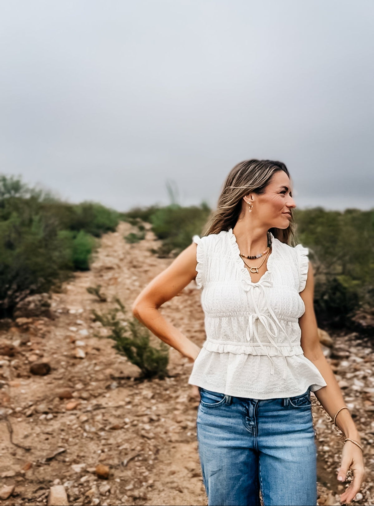 A woman wearing a white ruffled sleeveless blouse and blue jeans standing on a dirt path outdoors under an overcast sky.